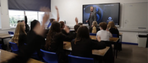 Image shows a class where the pupils are answering a question, hands in the air